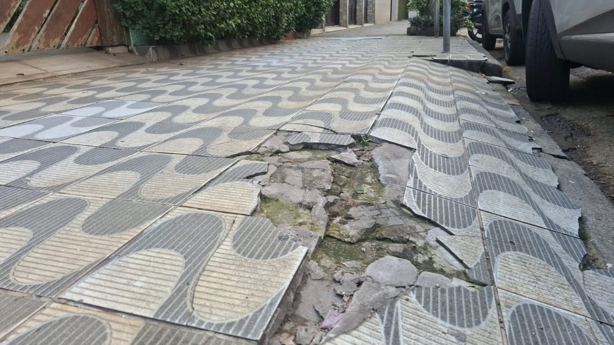 Foto em ângulo baixo e perspectiva profunda de uma calçada de Santos com padrão de ondas, mostrando danos extensos. Uma grande parte da calçada de azulejos foi removida ou quebrada, revelando um caminho profundo de concreto irregular e quebrado. As bordas restantes dos azulejos estão soltas. Carros estacionados estão à direita.