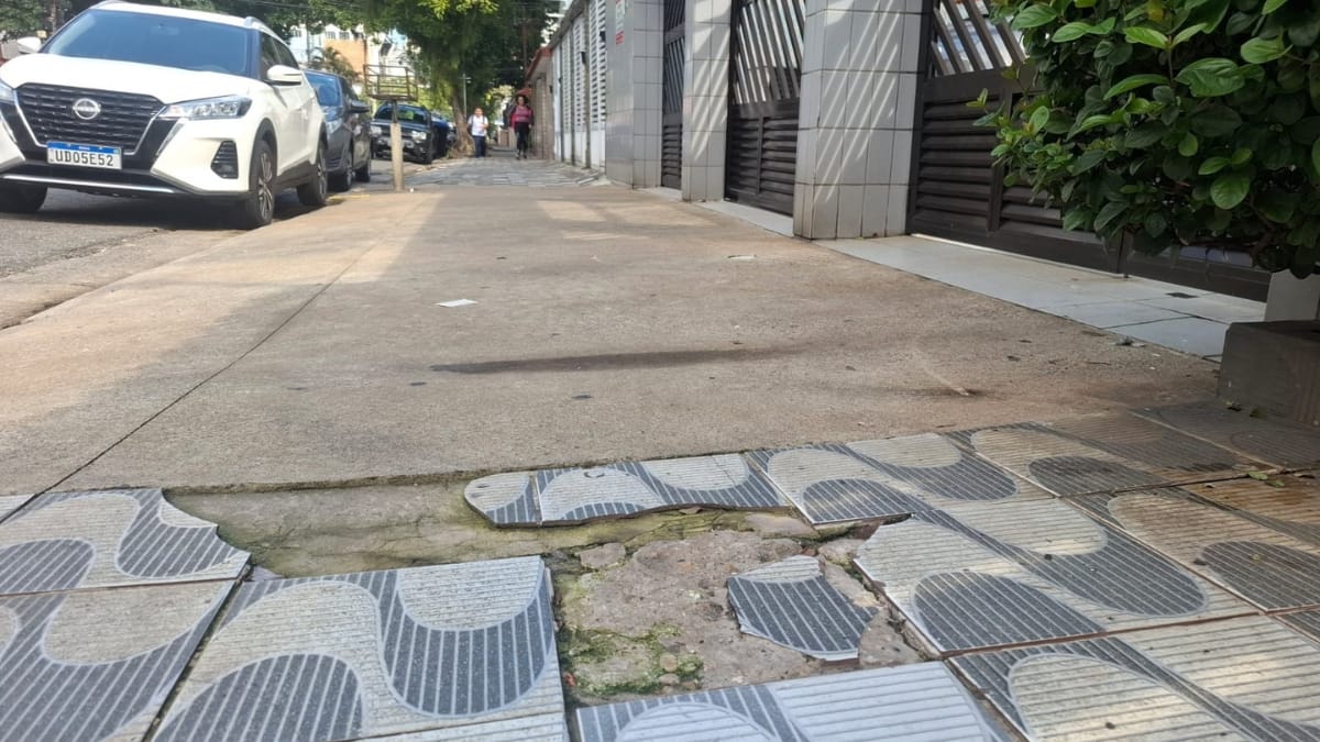 Foto em ângulo médio de uma calçada em Santos. O padrão de ondas de azulejos está ausente em uma seção no primeiro plano, criando um buraco perigoso e irregular no concreto subjacente. O resto da calçada parece estar em melhores condições. Carros estacionados e uma rua estão ao fundo.