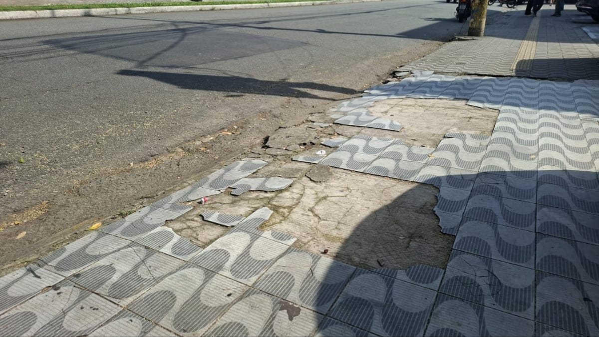 Foto em ângulo baixo de uma calçada de Santos com padrão de ondas. Uma área significativa está faltando azulejos, criando um grande buraco irregular que expõe concreto rachado e irregular abaixo. Mais azulejos soltos cercam o buraco. A rua está à esquerda.