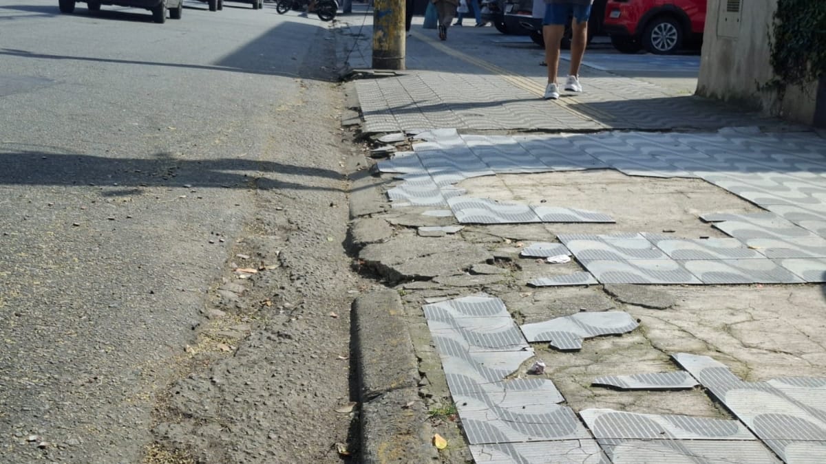 Uma foto focada em uma calçada urbana em Santos com padrão de ondas preto e branco. Uma grande seção de azulejos está faltando, revelando um buraco profundo e irregular na superfície de concreto inferior. Pernas de um pedestre caminham à direita.