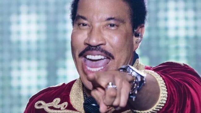 Lionel Richie se apresenta no palco e aponta para o público durante show ao vivo.