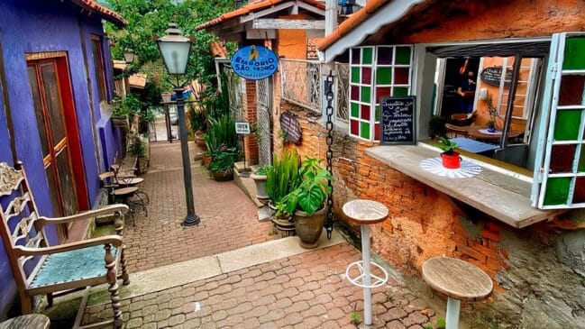 Uma charmosa ruela ou beco de paralelepípedos ladeado por construções de estilo rústico e colorido, com destaque para a fachada de tijolos aparentes do "Empório São Pedro" à direita