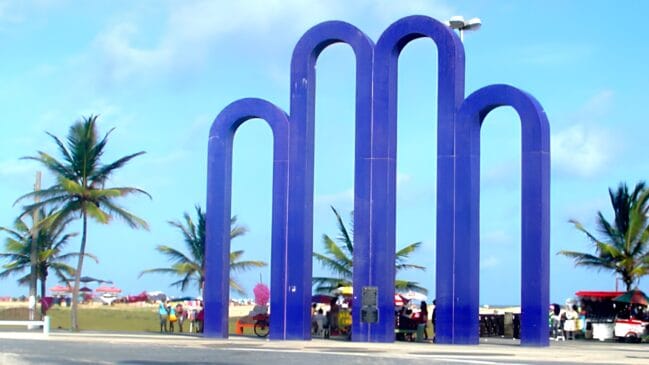 Monumento dos Arcos da Orla de Atalaia, em Aracaju, com coqueiros e praia ao fundo.