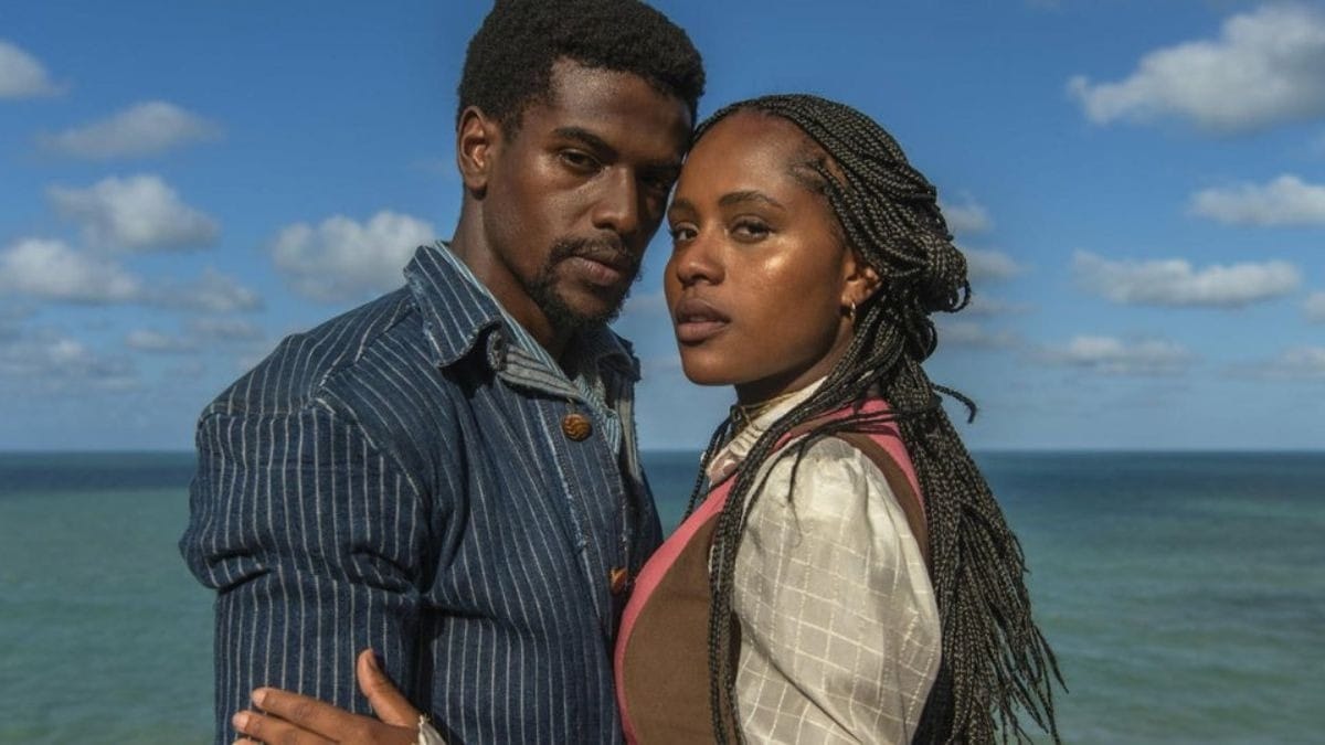 Um casal negro posa abraçado em um penhasco à beira-mar sob um céu azul com poucas nuvens, vestido com roupas de estilo rústico ou de época