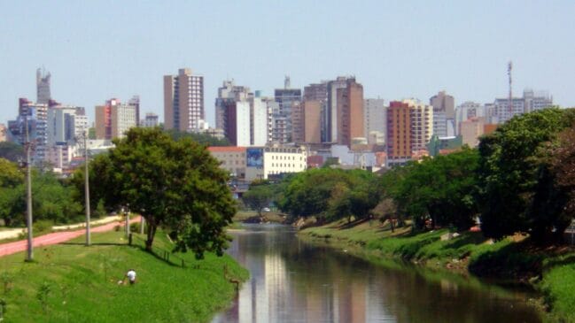 A imagem mostra uma paisagem urbana vista a partir de um rio ou canal, com bastante área verde em primeiro plano e um conjunto de edifícios altos ao fundo.