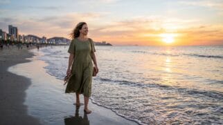 Mulher caminhando na areia molhada segurando as sandálias, com prédios da orla e o reflexo do sol no mar ao fundo.