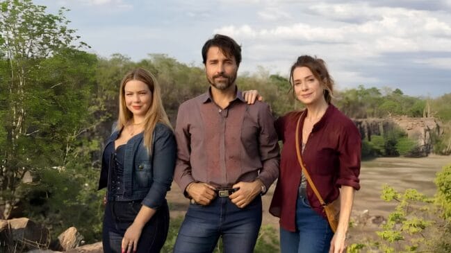 Três pessoas, um homem ao centro e duas mulheres nas laterais, posam para uma foto em um cenário de natureza ao ar livre, com árvores e formações rochosas ao fundo