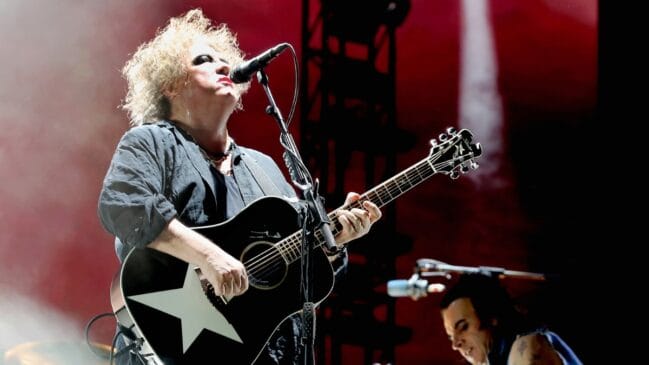 Robert Smith, vocalista da banda The Cure, cantando e tocando um violão preto com uma estrela branca no palco sob luzes vermelhas.