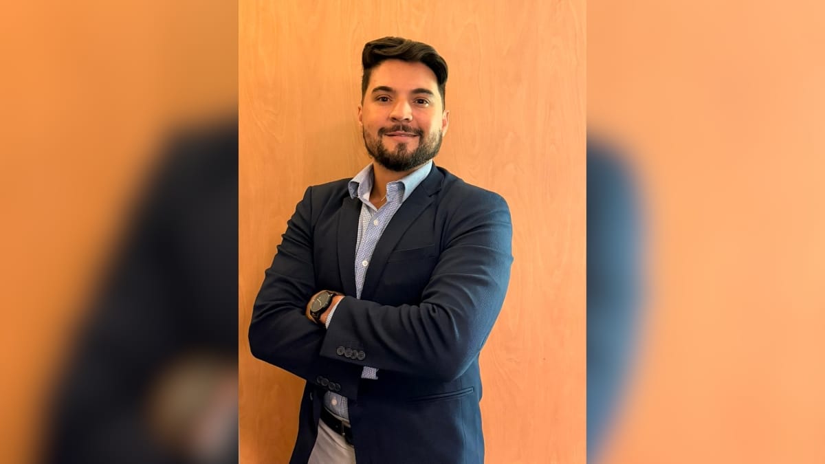 Homem jovem sorridente de barba e terno azul-marinho posa com os braços cruzados contra um fundo de madeira clara