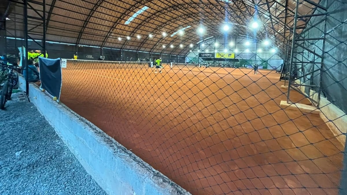 Foto em ângulo baixo mostrando a rede de proteção de metal e a quadra de saibro laranja vazia com as linhas brancas.