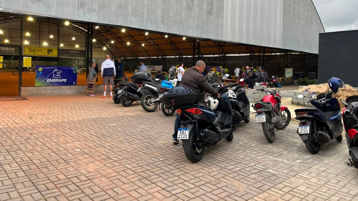 Estacionamento interno com motos enfileiradas e pessoas reunidas ao fundo sob a cobertura metálica das quadras.