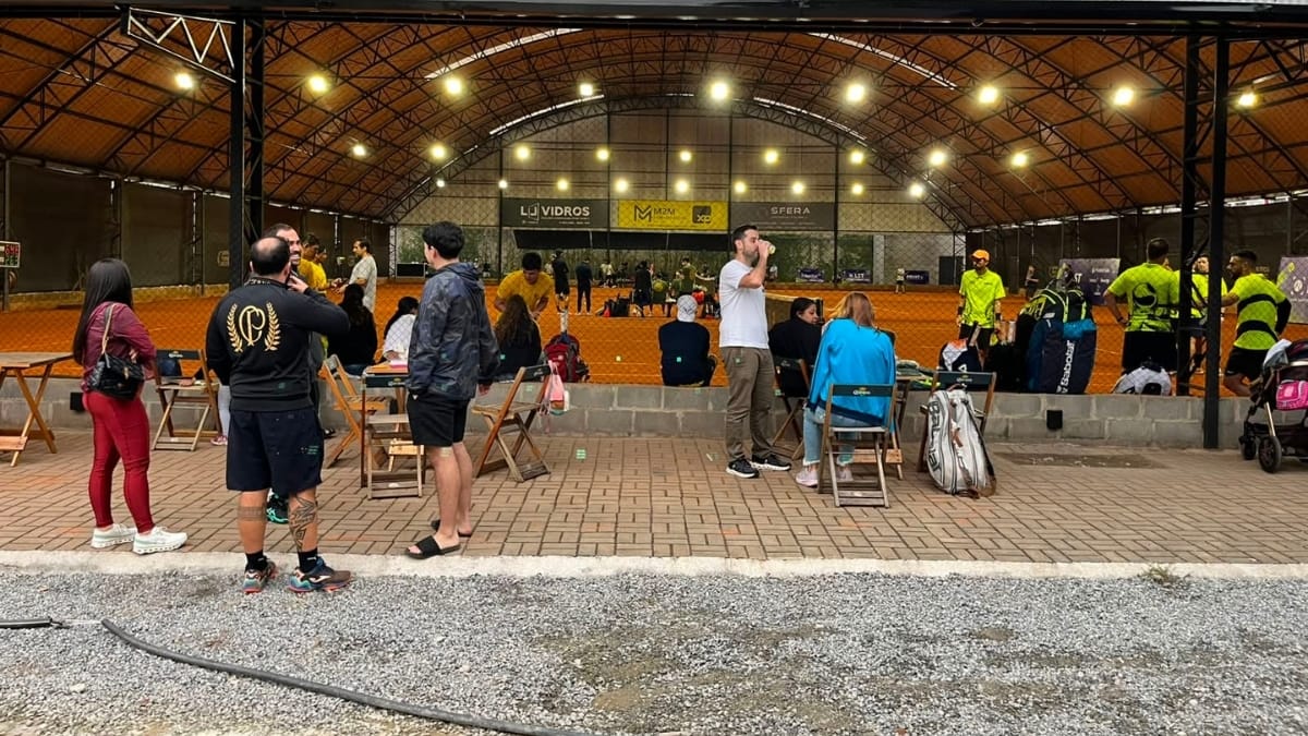 Público sentado em cadeiras de praia observando uma partida de tênis em uma quadra coberta e bem iluminada.