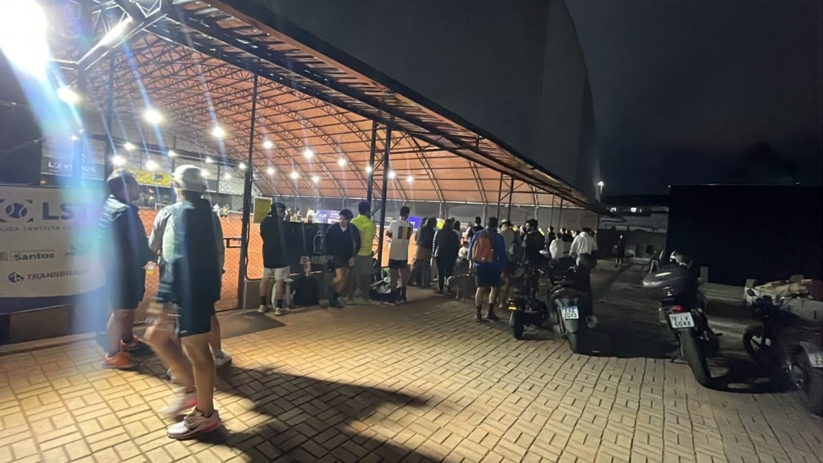 Vista externa do complexo esportivo à noite com pessoas caminhando em um pátio iluminado e motos estacionadas à direita.