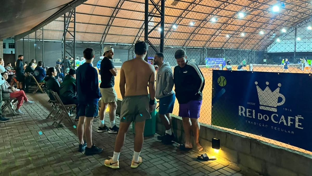 Grupo de homens em trajes esportivos conversando em um corredor interno de blocos de concreto ao lado de uma quadra de tênis.