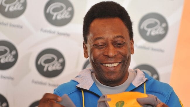 Fotografia em primeiro plano focando Pelé de jaqueta azul sorrindo com olhar direcionado para baixo enquanto manipula um objeto