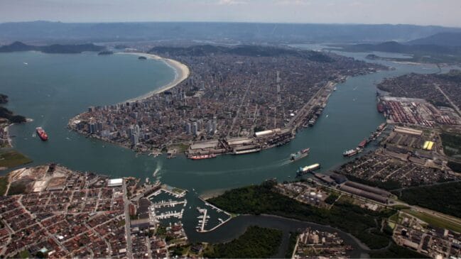 Fotografia aérea de plano aberto exibindo a vista panorâmica da cidade de Santos com o canal do porto e a orla da praia ao fundo