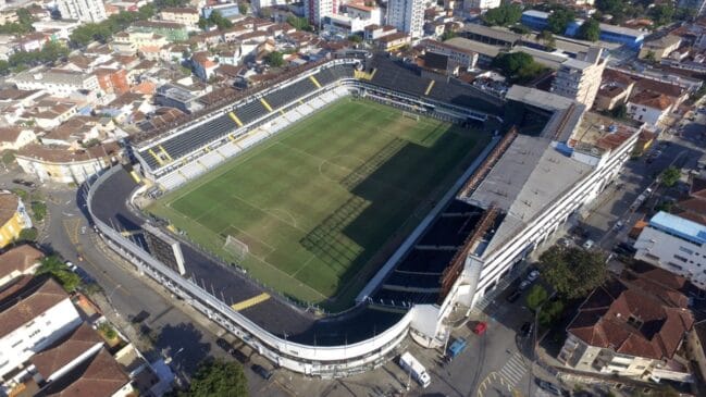 Fotografia aérea diagonal registrando toda a estrutura externa e o gramado do estádio da Vila Belmiro sob a luz do sol