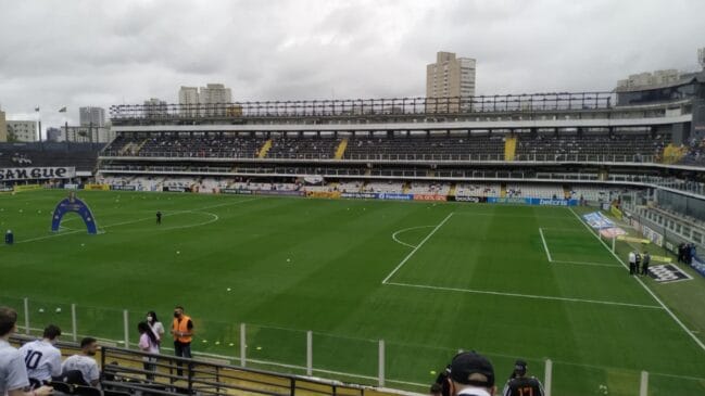 Fotografia em plano médio registrando o campo verde da Vila Belmiro a partir da arquibancada inferior durante os preparativos de um jogo