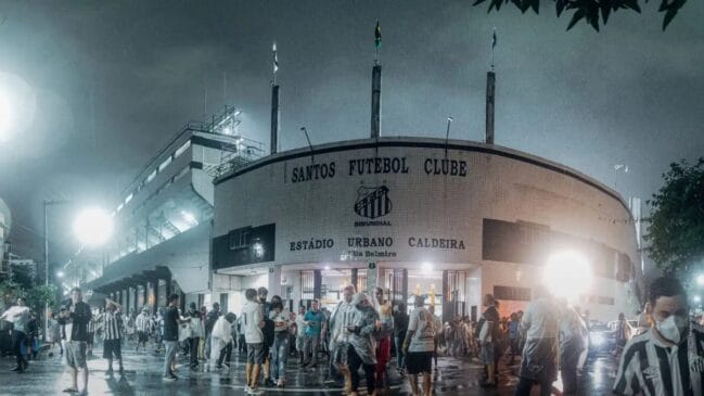 Fotografia horizontal registrando torcedores em frente à fachada principal do estádio da Vila Belmiro sob chuva leve à noite