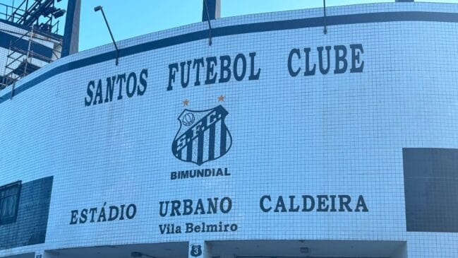 Fotografia em plano detalhe focando a parede de azulejos brancos com o escudo do Santos e o letreiro do Estádio Urbano Caldeira