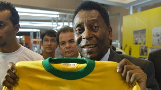 Fotografia em plano médio focando o ex-jogador Pelé sorrindo enquanto exibe uma camisa amarela de futebol com gola verde