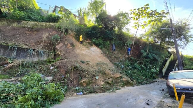 Encosta de morro caída com um carro estacionado e homens trabalhando na encosta do morro