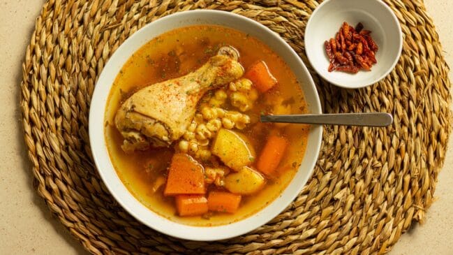 Fotografia horizontal em plano zenital de uma tigela de sopa de frango com legumes sobre um jogo americano de palha redonda ao lado de pimentas