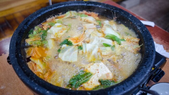 Fotografia horizontal de um cozido bósnio fervendo com folhas de repolho e pedaços de tofu dentro de uma panela de pedra escura