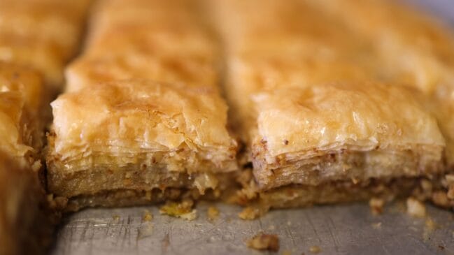 Fotografia horizontal em primeiro plano focando fatias quadradas de baklava folhada e dourada dispostas em uma assadeira