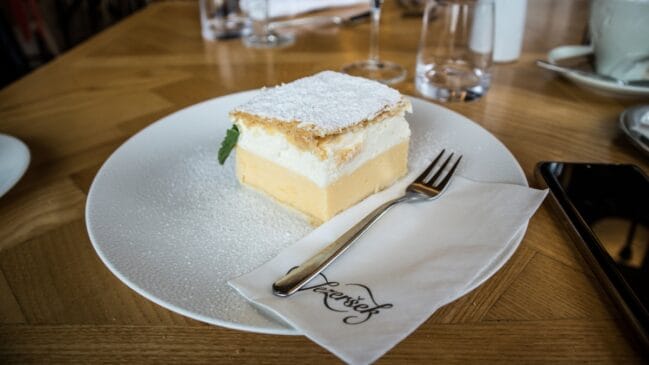 Fotografia horizontal de um pedaço quadrado de bolo cremsnita de baunilha e chantilly salpicado com açúcar de confeiteiro em um prato branco