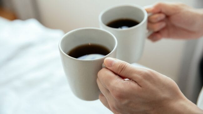 Fotografia horizontal de duas mãos segurando canecas brancas com café preto com fundo claro desfocado