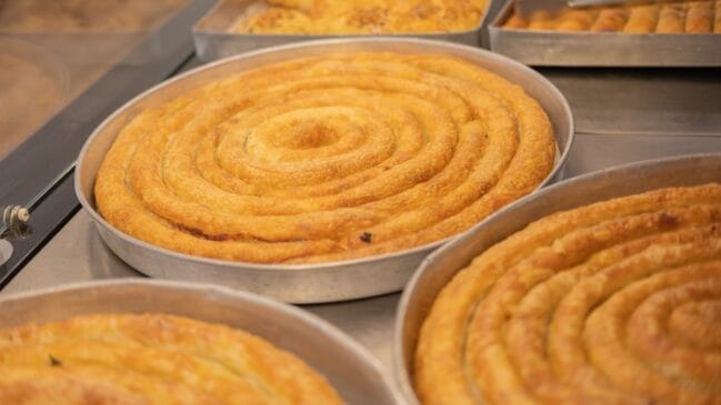 Fotografia horizontal de várias assadeiras redondas de metal contendo burek bósnio dourado em formato de espiral