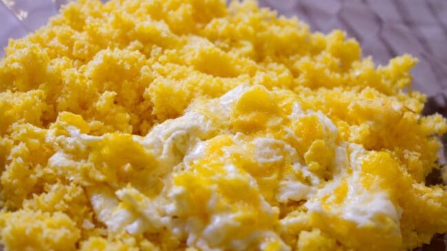 Fotografia horizontal com foco aproximado em uma porção de polenta amarela granulada servida com ovos mexidos por cima