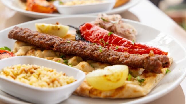 Fotografia horizontal de um prato com cevapi bósnio sobre pão folhado acompanhado de pimentão vermelho assado e arroz