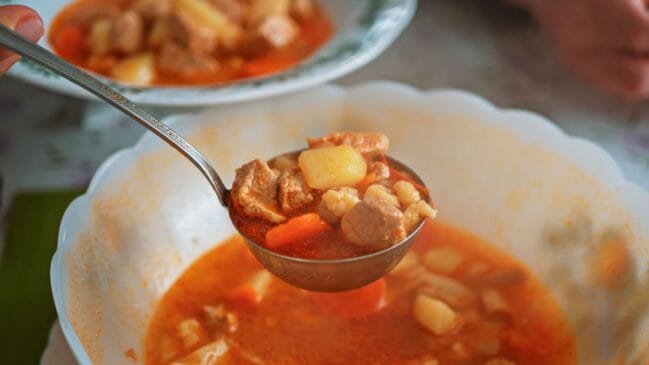 Fotografia horizontal mostrando uma concha de metal servindo um ensopado de carne com batatas e cenouras acima de uma tigela
