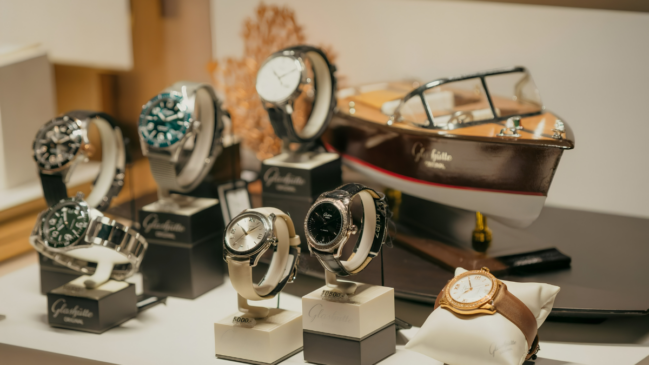 Imagem colorida, em plano médio e horizontal, mostrando uma vitrine iluminada com relógios de luxo da marca alemã Glashütte Original dispostos em suportes individuais, ao lado de uma miniatura de barco de madeira. No primeiro plano, alinhados sobre uma superfície branca, destacam-se três relógios: à esquerda, um modelo com pulseira metálica prateada e mostrador escuro; ao centro, um relógio com mostrador claro e pulseira de couro bege; e, à direita, um modelo com mostrador preto e pulseira de couro escura. Mais à direita, apoiado sobre uma pequena almofada branca, há um relógio de ouro com pulseira de couro marrom. Logo atrás, em suportes pretos ligeiramente mais altos, aparecem outros três relógios com caixas e mostradores em tons de preto, verde e branco. No canto superior direito, complementando a decoração da vitrine, há uma miniatura detalhada de um barco clássico de popa longa, feito de madeira envernizada escura e branca, que traz a inscrição dourada "Glashütte Original" em sua lateral. O fundo da imagem apresenta tons claros e desfocados.