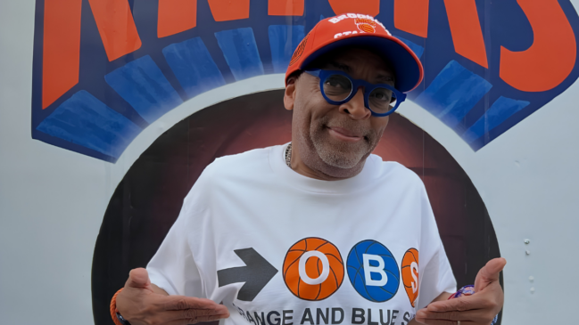 Uma fotografia colorida em plano médio apresenta o cineasta Spike Lee de pé, olhando diretamente para a câmera, com uma expressão ligeiramente divertida e acolhedora. Ele está de frente, com os dois braços dobrados e as mãos, com os dedos indicador e médio estendidos e os polegares para baixo, apontando para si mesmo ou para a estampa da sua camiseta. Ele veste um boné de beisebol laranja com abas azuis, que tem o logotipo do New York Knicks e as letras bordadas em branco e azul. Ele usa óculos de grau redondos com armações azuis brilhantes e uma camiseta branca limpa com um gráfico grande que combina o logotipo de uma seta preta, bolas de basquete laranja e azul, as letras "O B S" e o texto em arco "ORANGE AND BLUE". Um anel de ouro é visível no dedo anelar esquerdo. O fundo é uma parede cinza-branca, com uma porção de uma grande e desfocada inscrição em azul e laranja do logotipo "KNICKS" visível na parte superior. A iluminação é natural e uniforme, e a imagem está em foco.