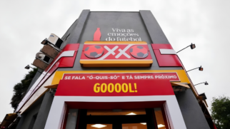 Foto em ângulo contra-plongée da fachada de uma loja de conveniência OXXO customizada para a Copa do Mundo. A estrutura cinza exibe no topo a frase "Viva as emoções do futebol" ao lado da silhueta de uma garrafa de Coca-Cola. Logo abaixo, o letreiro luminoso da marca traz duas bolas de futebol estilizadas ladeando as letras "XX". Na cobertura da entrada, uma faixa amarela exibe o texto "SE FALA ‘Ó-QUIS-SÓ’ E TÁ SEMPRE PRÓXIMO" e, logo abaixo, um grande painel vermelho destaca a palavra "GOOOOL!" em letras amarelas. O céu ao fundo está nublado.