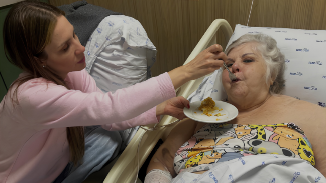 Uma mulher blusa rosa dando de comer para uma senhora que está deitada em uma maca