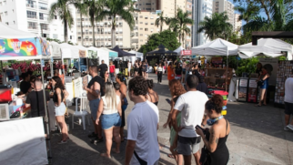 A imagem mostra uma feira de artesanato e gastronomia ao ar livre, realizada durante o dia em uma praça urbana pavimentada, cercada por palmeiras e edifícios residenciais altos ao fundo. Diversas barracas com tendas brancas enfileiram-se nas laterais, onde pessoas de diferentes idades caminham, conversam e observam os produtos. No primeiro plano, visitantes de costas e de perfil vestem roupas casuais de verão, como camisetas, regatas e shorts, sob a luz natural do sol que projeta sombras nítidas no chão.