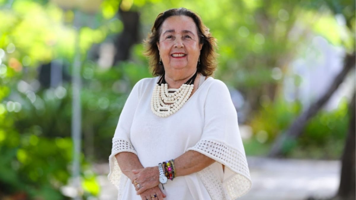 Mulher idosa sorridente, vestindo blusa branca e colar de contas, posando para foto após defender seu TCC, celebrando a educação na terceira idade.