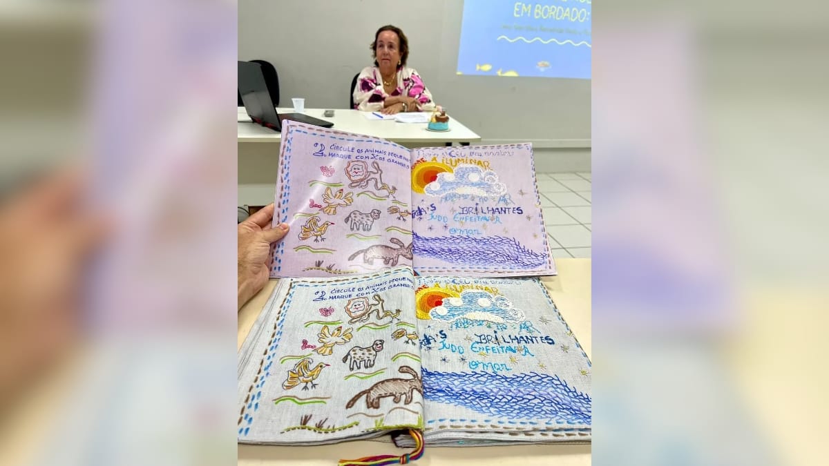 Trabalho de conclusão de curso apresentado como um livro artesanal bordado à mão, com ilustrações detalhadas de animais e elementos naturais, com a autora ao fundo na mesa de defesa.