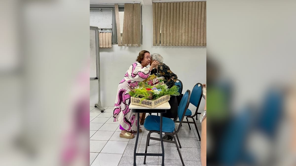 Formanda beijando sua mãe idosa, sentada à mesa, com uma cesta de flores entre elas em uma sala de aula, celebrando a conclusão do TCC com afeto e gratidão.