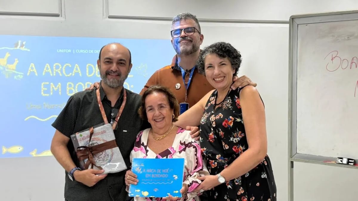 Autora de TCC sorridente segurando seu livro artesanal bordado, acompanhada por professores da banca examinadora em um ambiente acadêmico, celebrando a conclusão do projeto.