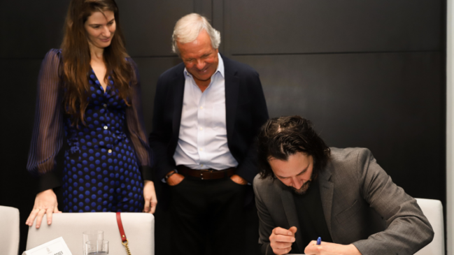 Keanu Reeves assinando um documento com duas pessoas olhando