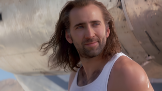 A imagem traz um plano médio do ator Nicolas Cage em sua juventude, caracterizado como o personagem Cameron Poe do filme Con Air (1997). Ele está ligeiramente de perfil, voltado para a direita, com um leve sorriso sereno no rosto e os olhos azuis direcionados para o horizonte. O ator tem cabelos castanhos longos, que caem desalinhados até a altura dos ombros e dão a impressão de estarem levemente ao vento. Sua pele é clara, e ele exibe uma barba rala por fazer em tom castanho-escuro. Ele veste uma regata branca simples de algodão com decote cavado, que deixa à mostra pelos no peito e seus ombros robustos. O fundo da cena é composto pela fuselagem texturizada e metálica de uma aeronave antiga, em tons claros e beges esmaecidos, que ocupa quase toda a área traseira, mantendo o foco principal na expressão do ator.
