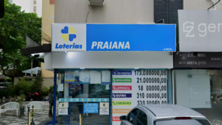 Descrição da Imagem (Acessibilidade — Para Cego Ver) A imagem captura a fachada da lotérica Praiana, localizada em uma área urbana. A composição destaca a identidade visual do estabelecimento e as informações sobre os prêmios acumulados. Elementos Principais Fachada e Letreiro: No topo, um grande painel horizontal exibe, à esquerda, o logotipo das Loterias Caixa (um trevo de quatro folhas nas cores azul e amarelo) seguido da palavra "Loterias" em azul. À direita, em letras brancas maiores sobre um fundo azul, destaca-se o nome "PRAIANA". Painel de Prêmios: À direita da entrada, um cartaz vertical lista os valores estimados para os próximos sorteios: +Milionária: 173.000.000,00 Mega-Sena: 21.000.000,00 Lotofácil: 1.800.000,00 Quina: 3.100.000,00 Timemania: 3.300.000,00 Entrada e Vitrine: A entrada possui estrutura de vidro, permitindo ver o interior do estabelecimento. Na parte inferior do vidro, há adesivos informativos azuis e amarelos, incluindo avisos de "Exclusivo para clientes". Elementos Secundários e Ambiente Entorno: À esquerda, nota-se uma área externa com piso de mosaico português (preto e branco), cadeiras de plástico brancas e uma motocicleta vermelha estacionada ao fundo. Uma pessoa aparece de costas, próxima à entrada. Edificação: À direita da lotérica, há uma parede cinza com um painel publicitário parcial onde se lê "ger" e números de telefone com o código de área (13), indicando a localização na região da Baixada Santista. O prédio acima da loja possui janelas com persianas escuras.