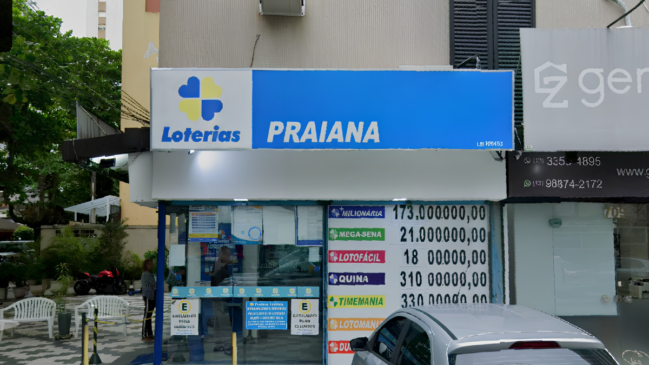 Descrição da Imagem (Acessibilidade — Para Cego Ver) A imagem captura a fachada da lotérica Praiana, localizada em uma área urbana. A composição destaca a identidade visual do estabelecimento e as informações sobre os prêmios acumulados. Elementos Principais Fachada e Letreiro: No topo, um grande painel horizontal exibe, à esquerda, o logotipo das Loterias Caixa (um trevo de quatro folhas nas cores azul e amarelo) seguido da palavra "Loterias" em azul. À direita, em letras brancas maiores sobre um fundo azul, destaca-se o nome "PRAIANA". Painel de Prêmios: À direita da entrada, um cartaz vertical lista os valores estimados para os próximos sorteios: +Milionária: 173.000.000,00 Mega-Sena: 21.000.000,00 Lotofácil: 1.800.000,00 Quina: 3.100.000,00 Timemania: 3.300.000,00 Entrada e Vitrine: A entrada possui estrutura de vidro, permitindo ver o interior do estabelecimento. Na parte inferior do vidro, há adesivos informativos azuis e amarelos, incluindo avisos de "Exclusivo para clientes". Elementos Secundários e Ambiente Entorno: À esquerda, nota-se uma área externa com piso de mosaico português (preto e branco), cadeiras de plástico brancas e uma motocicleta vermelha estacionada ao fundo. Uma pessoa aparece de costas, próxima à entrada. Edificação: À direita da lotérica, há uma parede cinza com um painel publicitário parcial onde se lê "ger" e números de telefone com o código de área (13), indicando a localização na região da Baixada Santista. O prédio acima da loja possui janelas com persianas escuras.