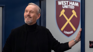 A imagem mostra o ator Anthony Head caracterizado como seu personagem Rupert Mannion na série Ted Lasso. Ele é um homem branco de meia-idade, com cabelos curtos e grisalhos penteados para o lado, e uma barba rala também grisalha concentrada ao redor da boca e no queixo. Ele está posicionado do lado esquerdo da imagem, de perfil três quartos voltado para a direita, exibindo um sorriso sutil e olhando para cima com uma expressão confiante. O ator veste uma blusa de gola alta preta sob uma jaqueta escura aberta. Seu braço esquerdo está estendido para o lado direito, com a mão apoiada no batente de uma porta azul. No pulso esquerdo, ele usa um relógio de metal prateado. Ao fundo, através da abertura da porta, destaca-se na parede cinza o escudo oficial do time de futebol inglês West Ham United. O emblema tem formato de escudo heráldico nas cores grená e azul, com dois martelos amarelos cruzados no centro e as inscrições "WEST HAM UNITED" no topo e "LONDON" na base.