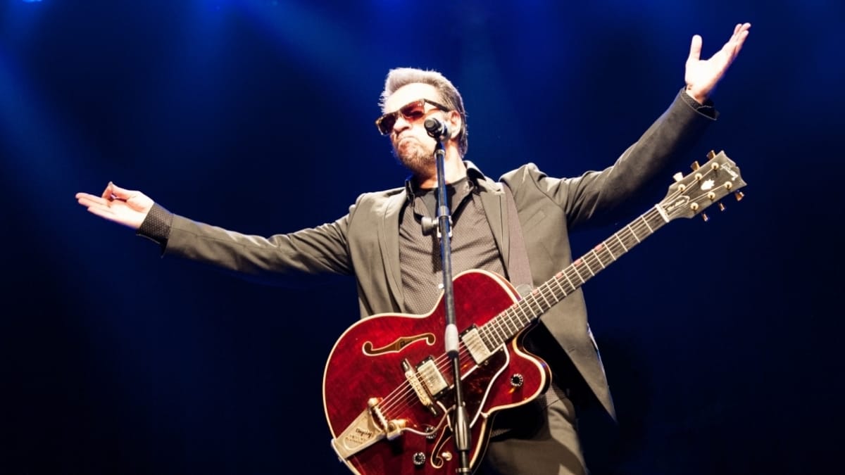 Cantor Marcelo Nova no palco, usando óculos escuros e tocando guitarra elétrica vermelha durante show com iluminação azul.
